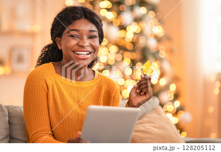 Online shopping at home during covid-19 self-isolation. Millennial african american laughing lady holds tablet and credit card for shopping in living room interior with Xmas tree and glowing garlands Online shopping at home during covid-19 self-isolation. Millennial african american laughing lady holds tablet and credit card for shopping in living room interior with Xmas tree and glowing garlands 128205112