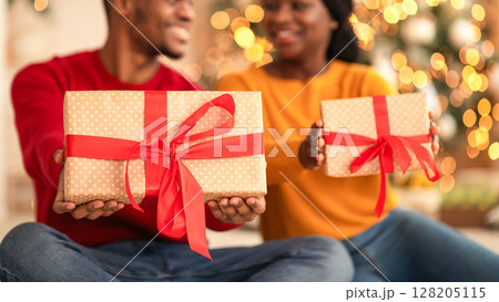 Gifts and New Year mood in coronavirus pandemic. Happy millennial african american man and woman show boxes with ribbons and look at each other in living room interior with Xmas tree with garlands Gifts and New Year mood in coronavirus pandemic. Happy millennial african american man and woman show boxes with ribbons and look at each other in living room interior with Xmas tree with garlands 128205115