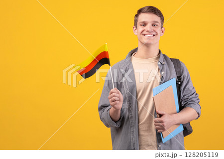 Modern education, language learning and knowledge exchange. Teen cheerful guy in glasses with backpack, notebooks and small Germany flag, isolated on orange background, copy space, studio shot 128205119