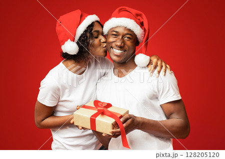 Happy black woman giving her boyfriend Christmas gift on orange studio background, copy space. Cheerful african american family in Santa hats celebrating New Year together, lady kissing man 128205120