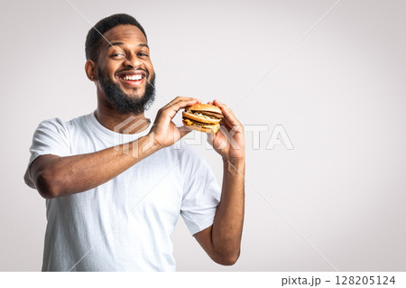 Smiling Black Guy Holding Unhealthy Burger Posing Standing On White Background, Looking At Camera. Male Nutrition And Fast Food Overeating Habit, Cheat Meal On A Diet Concept. Studio Shot Smiling Black Guy Holding Unhealthy Burger Posing Standing On White Background, Looking At Camera. Male Nutrition And Fast Food Overeating Habit, Cheat Meal On A Diet Concept. Studio Shot 128205124