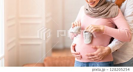 Muslim Husband Holding Tiny Shoes Hugging Pregnant Wife's Belly Hugging Standing At Home. Family Happiness And New Life. Happy Parents-To-Be Awaiting Baby Concept. Free Space For Text 128205254