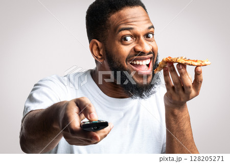 Black Man Eating Pizza And Watching TV Pointing Remote Control To Camera Posing Over White Studio Background. Junk Food Eater Watches Television Enjoying Unhealthy Fastfood. Overeating Nutrition Habit 128205271