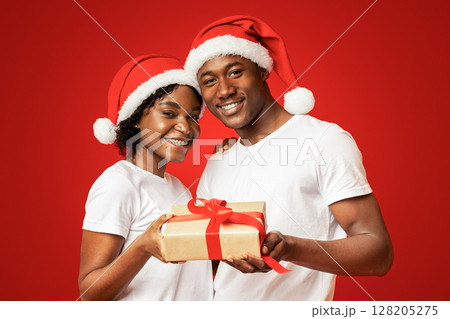 Cheerful african american couple celebrating New Year together, wearing Santa hats, embracing, holding gift box and smiling at camera over orange studio background, copy space. Winter holidays concept Cheerful african american couple celebrating New Year together, wearing Santa hats, embracing, holding gift box and smiling at camera over orange studio background, copy space. Winter holidays concept 128205275