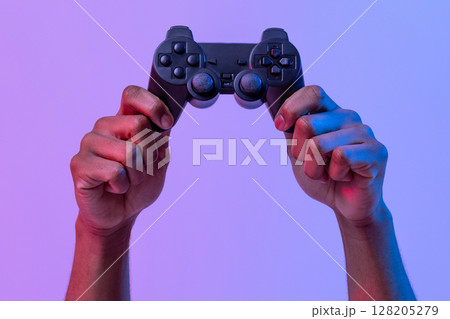 Black male hands holding joystick for video games over purple background with neon lighting, unrecognizable man enjoying modern technologies for gaming, cropped image, panorama with copy space 128205279