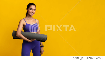 Wellness. Smiling millennial female athlete holding yoga mat standing isolated on orange studio background. Positive young woman promoting healthy lifestyle, ready for her morning practice 128205288