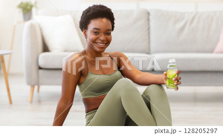 Stay Hydrated. African Fit Woman Holding Bottle Of Infused Water Drinking During Workout Sitting On Gymnastics Mat Posing At Home, Smiling To Camera. Healthy Hydration And Weight Loss Concept 128205297