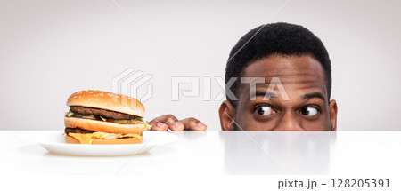 Funny Hungry African Guy Looking At Tasty Burger On Desk Having Food Craving Posing In Studio On White Background. Overeating Habit, Dieting And Nutrition, Cheat Meal Concept. Panorama 128205391