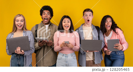 Gossip, shocking news, study with online technology and teamwork. Surprised teens multiracial students learning, using digital gadgets, isolated on orange background, copy space, studio shot 128205465
