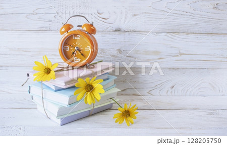 Alarm clock and stack of books with fresh flowers between pages, as bookmarks. On white wooden background, concept back to school, education. Alarm clock and stack of books with fresh flowers between pages, as bookmarks. On white wooden background, concept back to school, education. 128205750