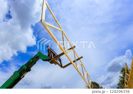 Forklift telehandler machinery raises wooden trusses beams high in air as fluffy clouds fill blue sky during workday. 128206356
