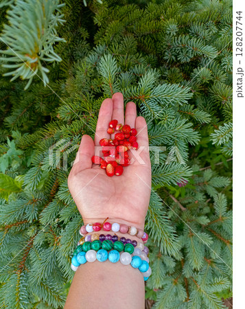 A hand with bracelets in a gentle pose cupping wild strawberries against lush greenery representing a bohemian summer 128207744