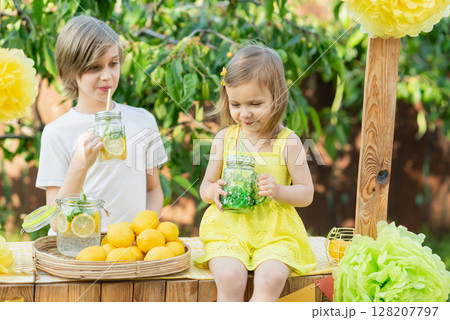 Children drink natural lemonade at stand in park. Summer refreshing natural drink lemonade. Detox fruit infused flavored water, cocktail in a beverage dispenser with fresh fruits Children drink natural lemonade at stand in park. Summer refreshing natural drink lemonade. Detox fruit infused flavored water, cocktail in a beverage dispenser with fresh fruits 128207797