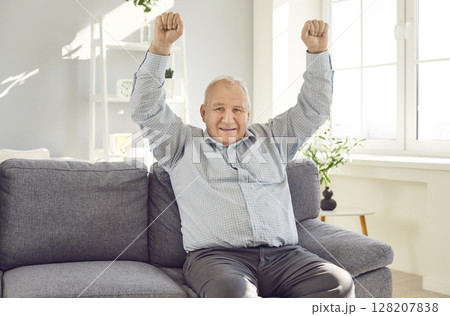 Elderly excited senior man sitting on sofa at home holding hands up in fist like a winner Elderly excited senior man sitting on sofa at home holding hands up in fist like a winner 128207838