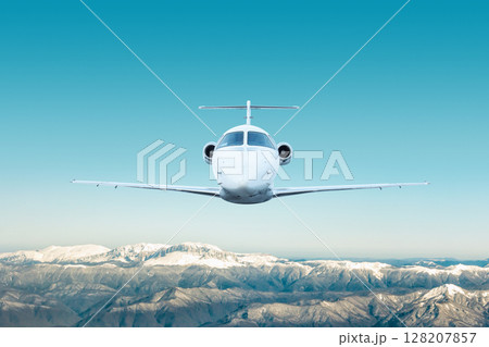 Front view of the white modern luxury executive aircraft flies over snow covered mountains 128207857