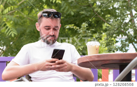 Bearded man wearing sunglasses and a white t shirt, sipping iced coffee at an outdoor cafe, using a smartphone to browse social media while enjoying a sunny summer day 128208508