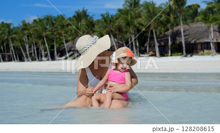 Mom sits in clear ocean water with baby daughter during tropical vacation Mom sits in clear ocean water with baby daughter during tropical vacation 128208615