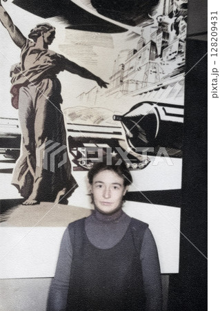 Vintage portrait of a young woman near the USSR poster Motherland Calls in the club of the contingent of Soviet troops in the GDR. Retro photo 1970. Vintage portrait of a young woman near the USSR poster Motherland Calls in the club of the contingent of Soviet troops in the GDR. Retro photo 1970. 128209431
