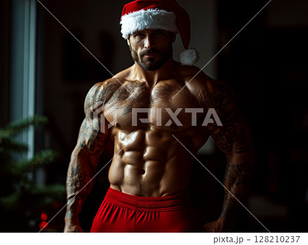 A muscular man wearing red shorts and a Santa hat stands confidently in a dimly lit room. Concept of holiday fitness. For fitness-themed holiday content. 128210237