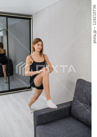Young woman doing lunge stretch with foot on sofa. 128210736