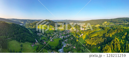 This aerial view of Tanvaldsky Spicak in the Jizera Mountains highlights green hills, dense forests, and charming villages under a clear blue sky in the morning light. 128211259