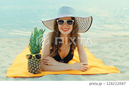 Summer vacation, happy smiling young woman with pineapple in hat with sunglasses lying on sea beach 128212087