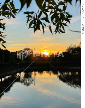 Beautiful sunset over pond in a city park. The sun goes down behind the trees. Clouds are reflected in the water. Vertical photography. Beautiful sunset over pond in a city park. The sun goes down behind the trees. Clouds are reflected in the water. Vertical photography. 128212547