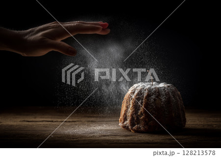 Hand Sprinkling Icing Sugar on Apple Bundt Cake on Old Wooden Table with Dark Background 128213578