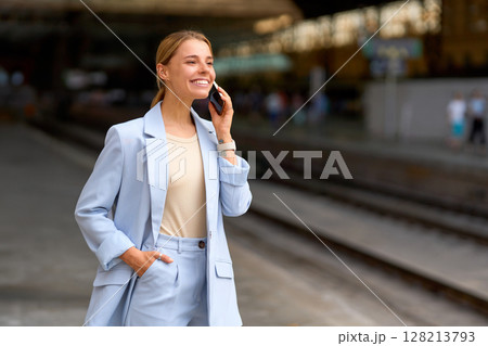 A confident woman is happily talking on her phone while standing at the busy train station 128213793