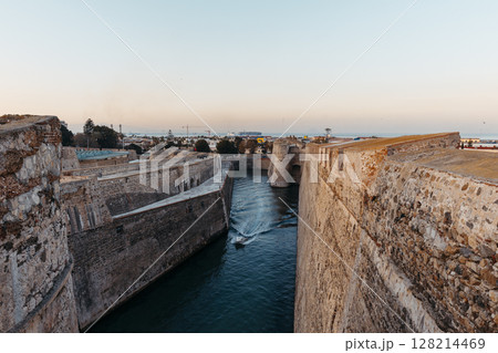 General view of the moat of the Royal Walls of Ceuta at sunset while a ship crosses it 128214469