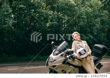 Stylish senior woman with gray hair and motorcycle posing on roadside, symbol of freedom and grace 128215454