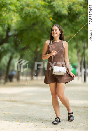 Pretty young woman in summer dress with handbag walking and posing in park on spring day 128215963