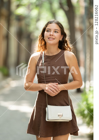 Beautiful smiling young lady wearing short dress walking shady tree lined street and relax in summer weather with sunshine outdoor Beautiful smiling young lady wearing short dress walking shady tree lined street and relax in summer weather with sunshine outdoor 128216062