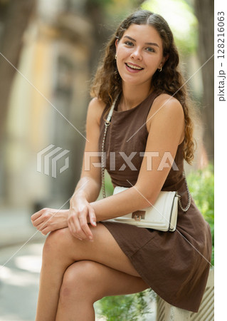 Beautiful young lady wearing dress walking shady street and sitting concrete flower bed with plants, taking break from heat of day Beautiful young lady wearing dress walking shady street and sitting concrete flower bed with plants, taking break from heat of day 128216063
