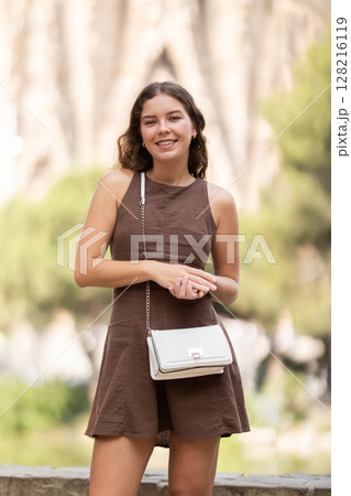 Pretty young woman in summer dress with handbag walking and posing in park on spring day 128216119