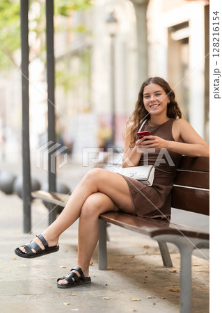 young woman sits on a bench in summer with a phone in her hands 128216154