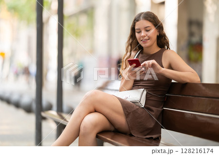young woman sits on a bench in summer with a phone in her hands 128216174