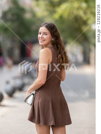 Back view of beautiful stylish young girl in dress with white handbag posing on city street on summer day Back view of beautiful stylish young girl in dress with white handbag posing on city street on summer day 128216216