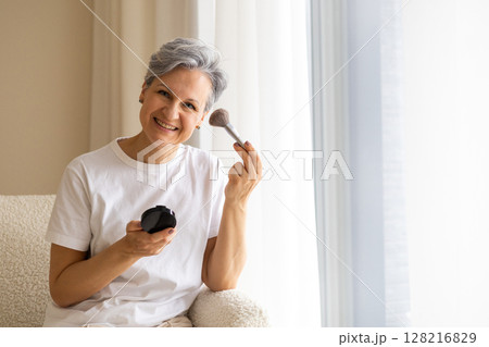 Beautiful smiling elderly woman pensioner during menopause with gray hair applies anti-aging makeup Beautiful smiling elderly woman pensioner during menopause with gray hair applies anti-aging makeup 128216829
