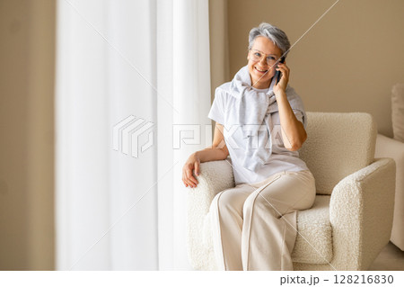 An elderly, mature, aged woman pensioner with gray hair sits in a chair near the window. An elderly, mature, aged woman pensioner with gray hair sits in a chair near the window. 128216830