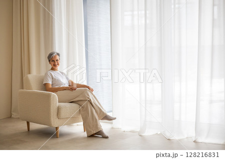 An elderly mature beautiful well-groomed woman pensioner sits near the window  128216831