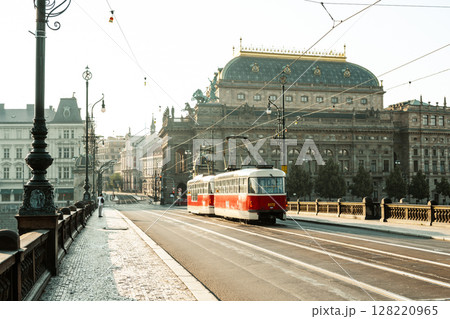 A vintage tram glides along the tracks past the National Theatre in Prague. The early morning light casts a warm glow on the historic architecture, creating a serene atmosphere. A vintage tram glides along the tracks past the National Theatre in Prague. The early morning light casts a warm glow on the historic architecture, creating a serene atmosphere. 128220965