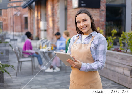 A smiling waitress in a restaurant holds a tablet, looking at the camera. Diners are in the background. A smiling waitress in a restaurant holds a tablet, looking at the camera. Diners are in the background. 128221288