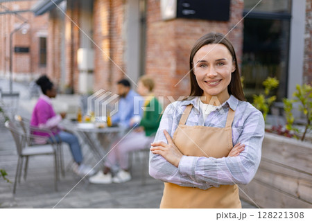 A smiling waitress in an apron poses in front of her outdoor restaurant, customers sit at tables in the background. A smiling waitress in an apron poses in front of her outdoor restaurant, customers sit at tables in the background. 128221308
