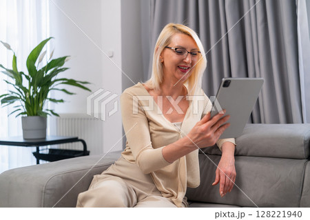 Blonde woman chatting with friend online using tablet on couch. Relaxed mature lady in casual outfit talking with her friend through video call, sitting comfortably at home. 128221940