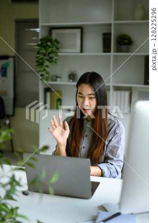Young woman smiling and waving during a video call at her home office Young woman smiling and waving during a video call at her home office 128225296