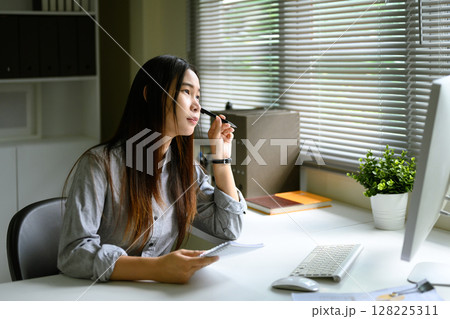Pensive businesswoman sitting at desk, gazing out the window with a pen in hand Pensive businesswoman sitting at desk, gazing out the window with a pen in hand 128225311