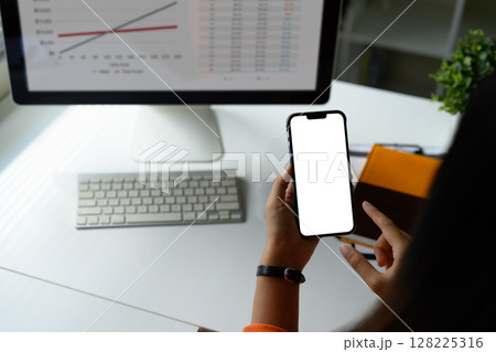 Hands holding smartphone with blank screen in front of desktop displaying financial graphs and data Hands holding smartphone with blank screen in front of desktop displaying financial graphs and data 128225316