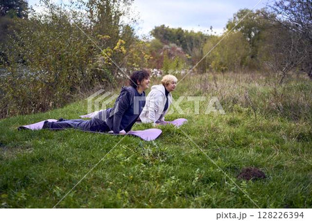 middle-aged woman teaches her old mother somatic exercises and yoga to maintain health by the river in autumn 128226394