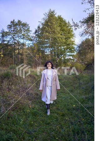 middle-aged woman with dark curly hair in light beige suit and coat enjoying nature in autumn forest 128226413